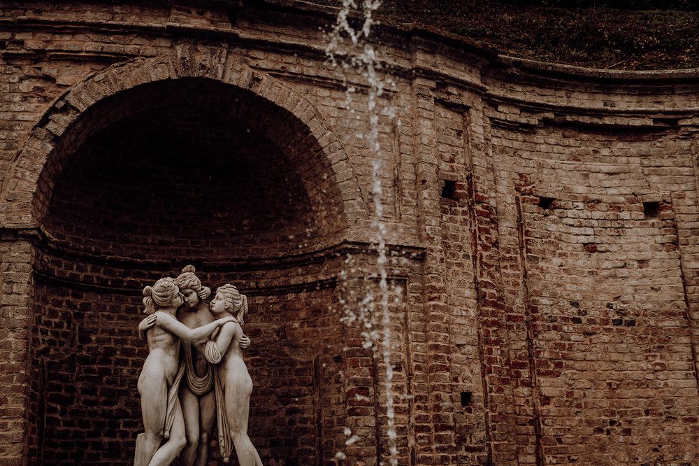 Classical stone fountain sculpture of three figures embracing, set against an aged brick wall with water cascading.