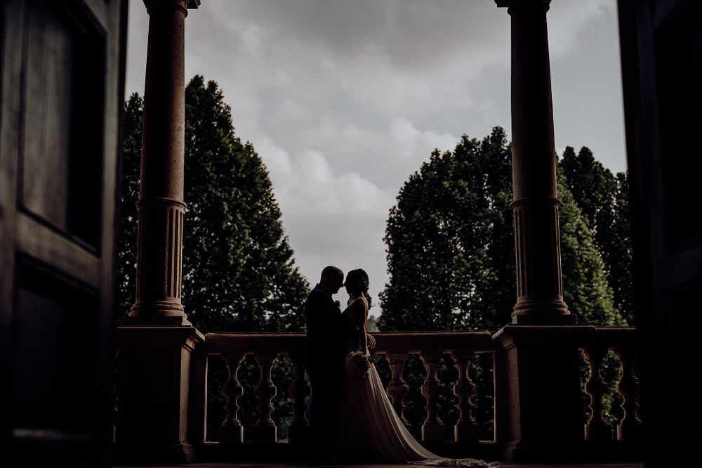 Romantic silhouette of bride and groom sharing a kiss on an elegant columned balcony under dramatic cloudy skies.