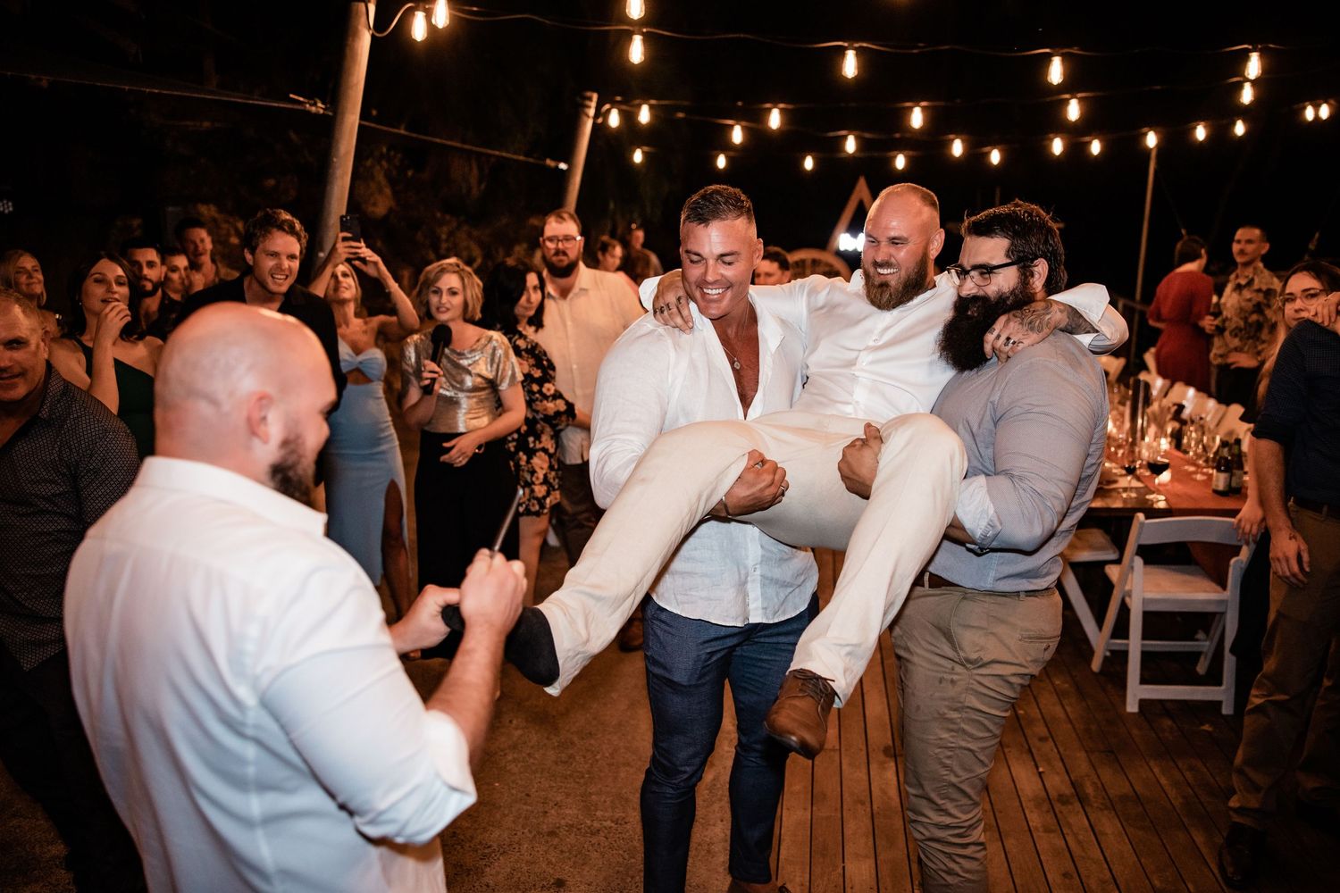Wedding guests lift and carry celebrating people during lively outdoor reception under string lights.