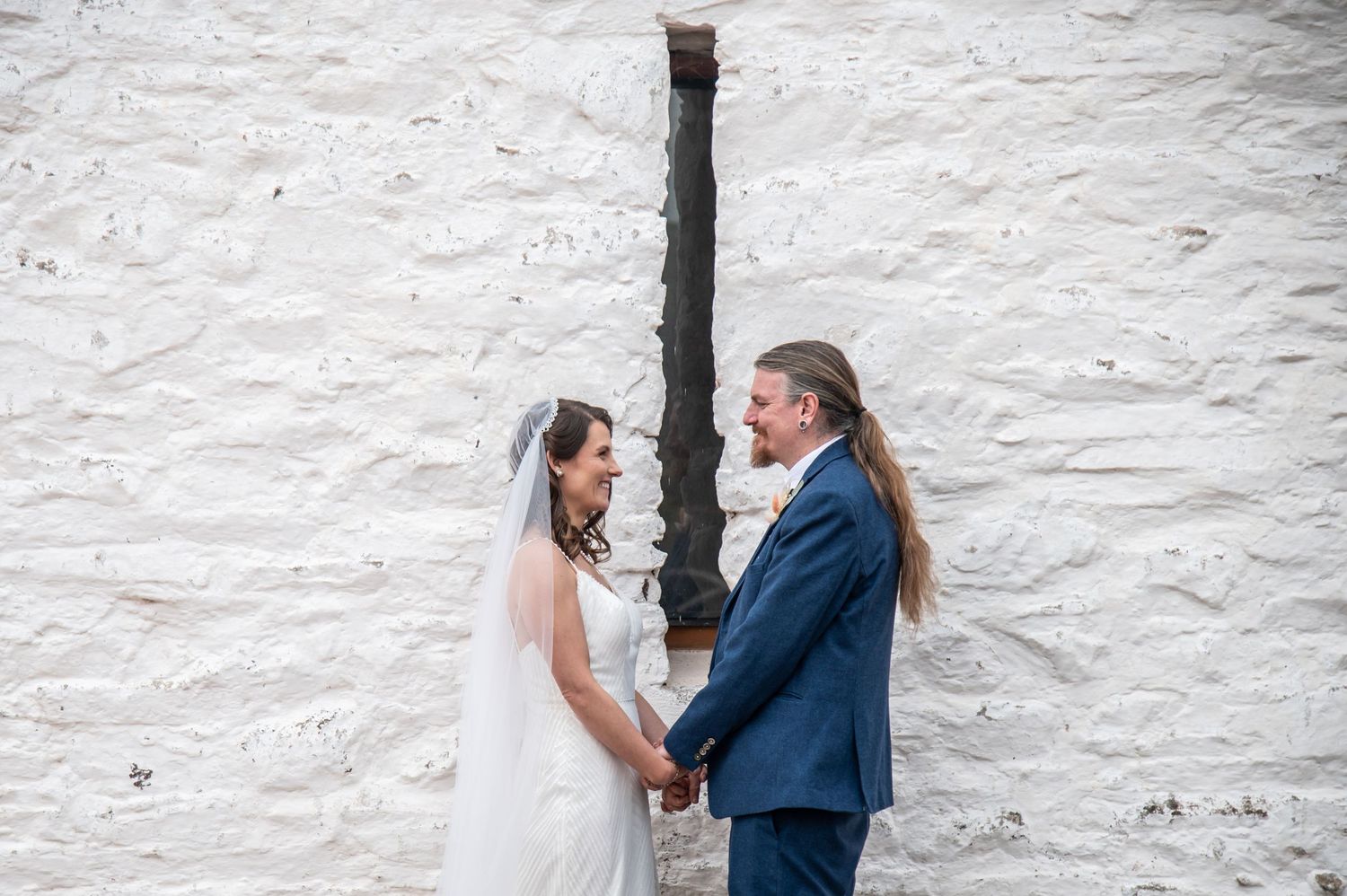 A couple poses against a white stone wall for wedding portraits.