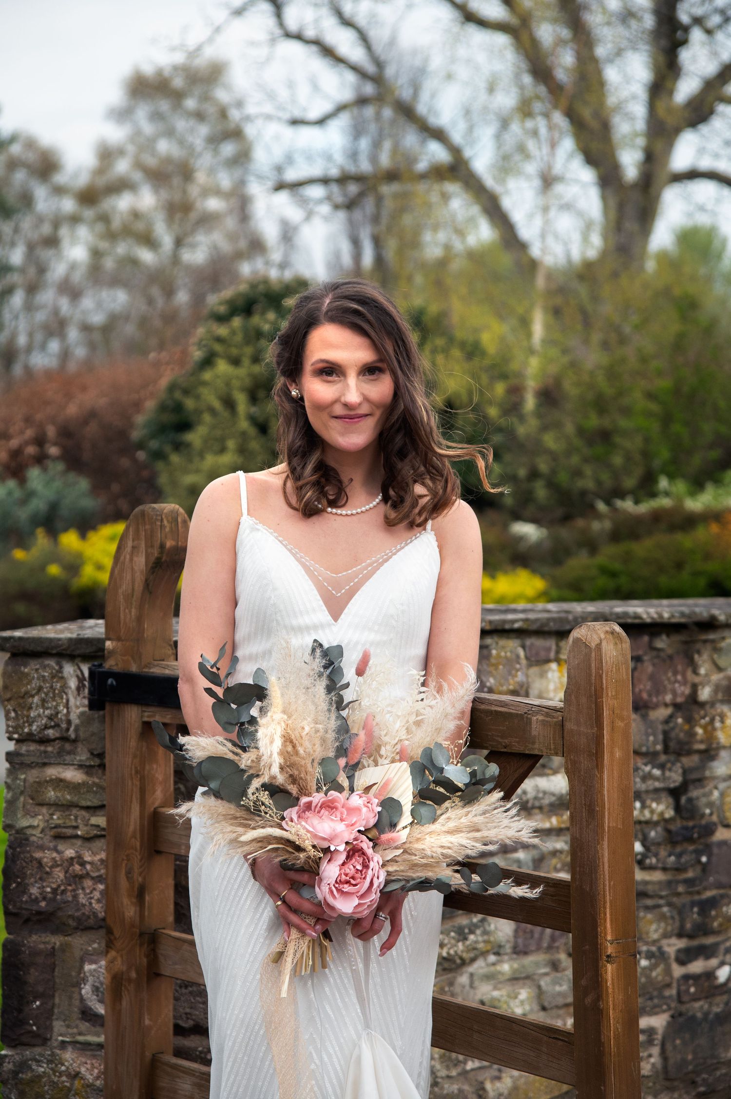A person in a white wedding dress holds a bouquet with dried flowers and pink roses by a stone wall.