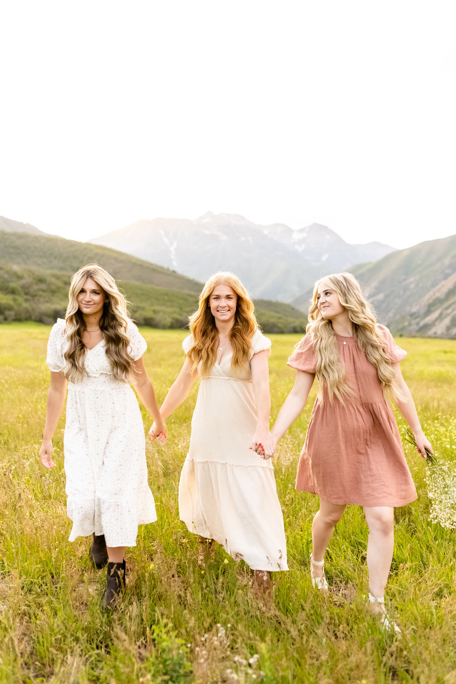 Three women in flowing dresses walk hand in hand through a mountain meadow.