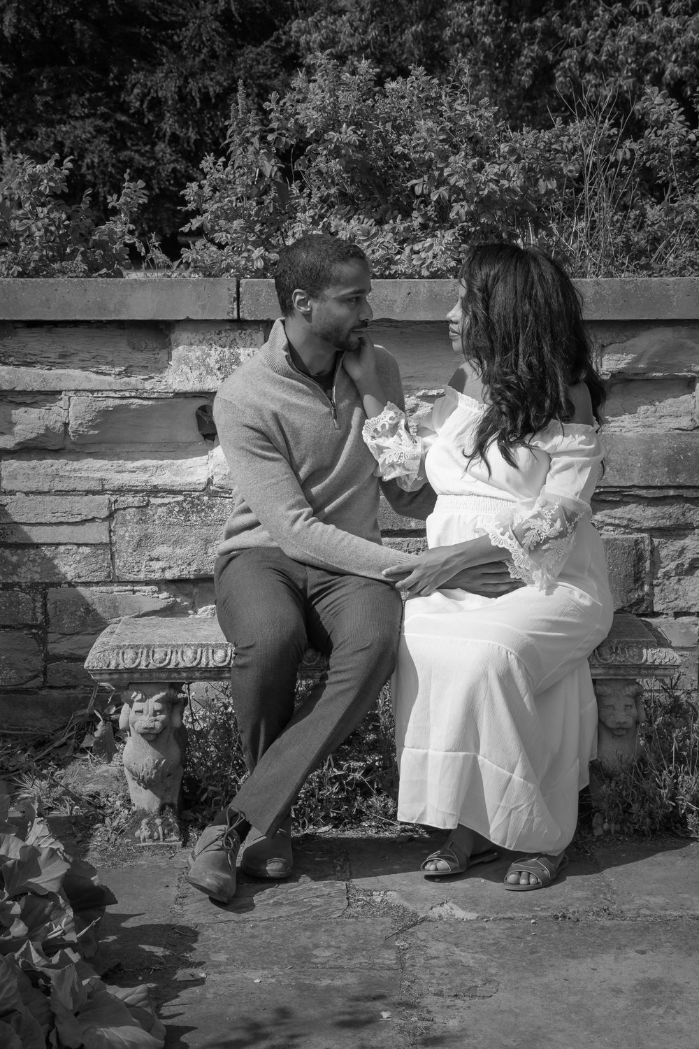 Black and white photo of expectant parents sitting together on stone wall surrounded by greenery.