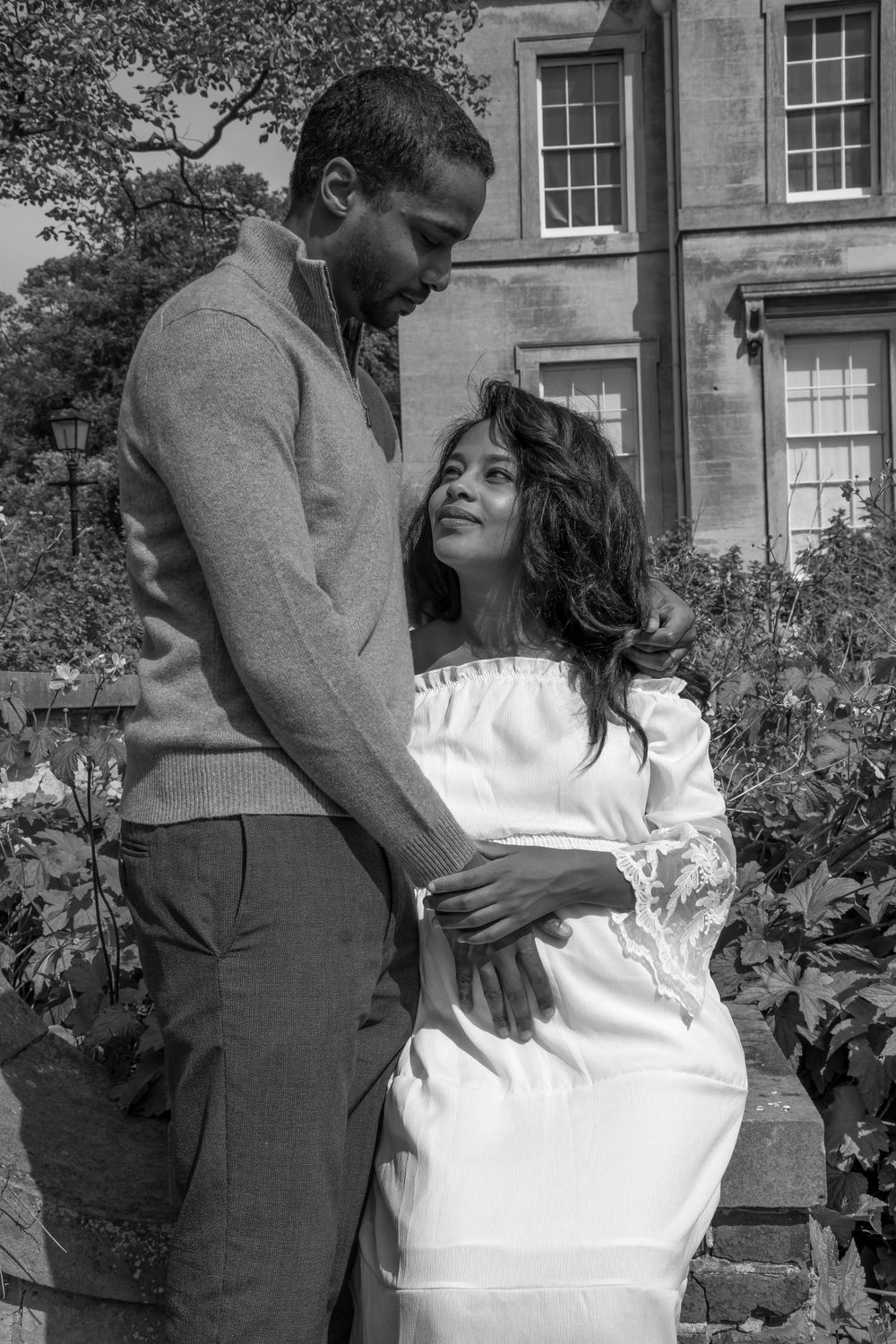 Intimate black and white maternity portrait with couple embracing against garden backdrop.