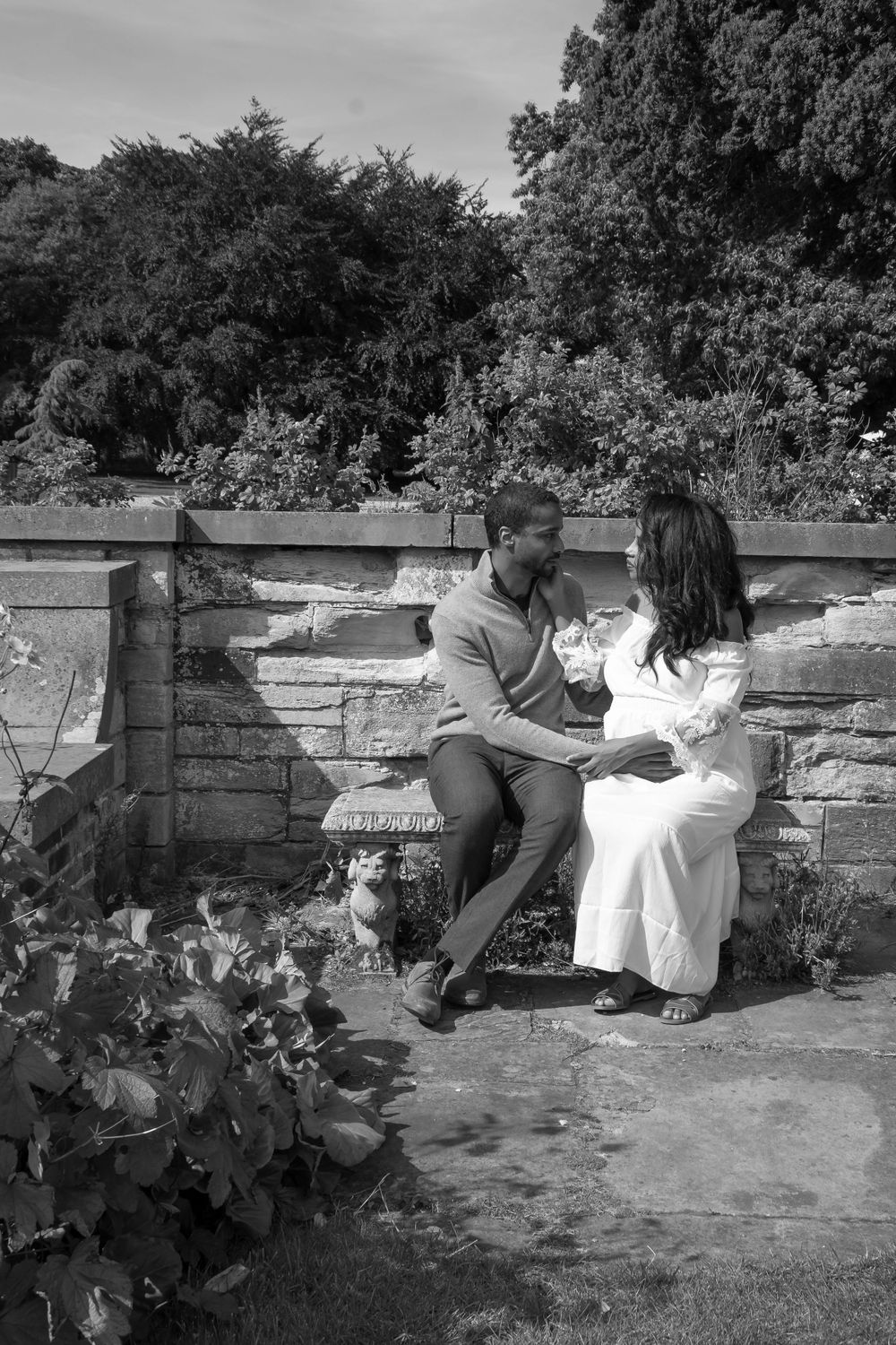 Expecting couple shares a tender moment while sitting on stone wall in garden setting.