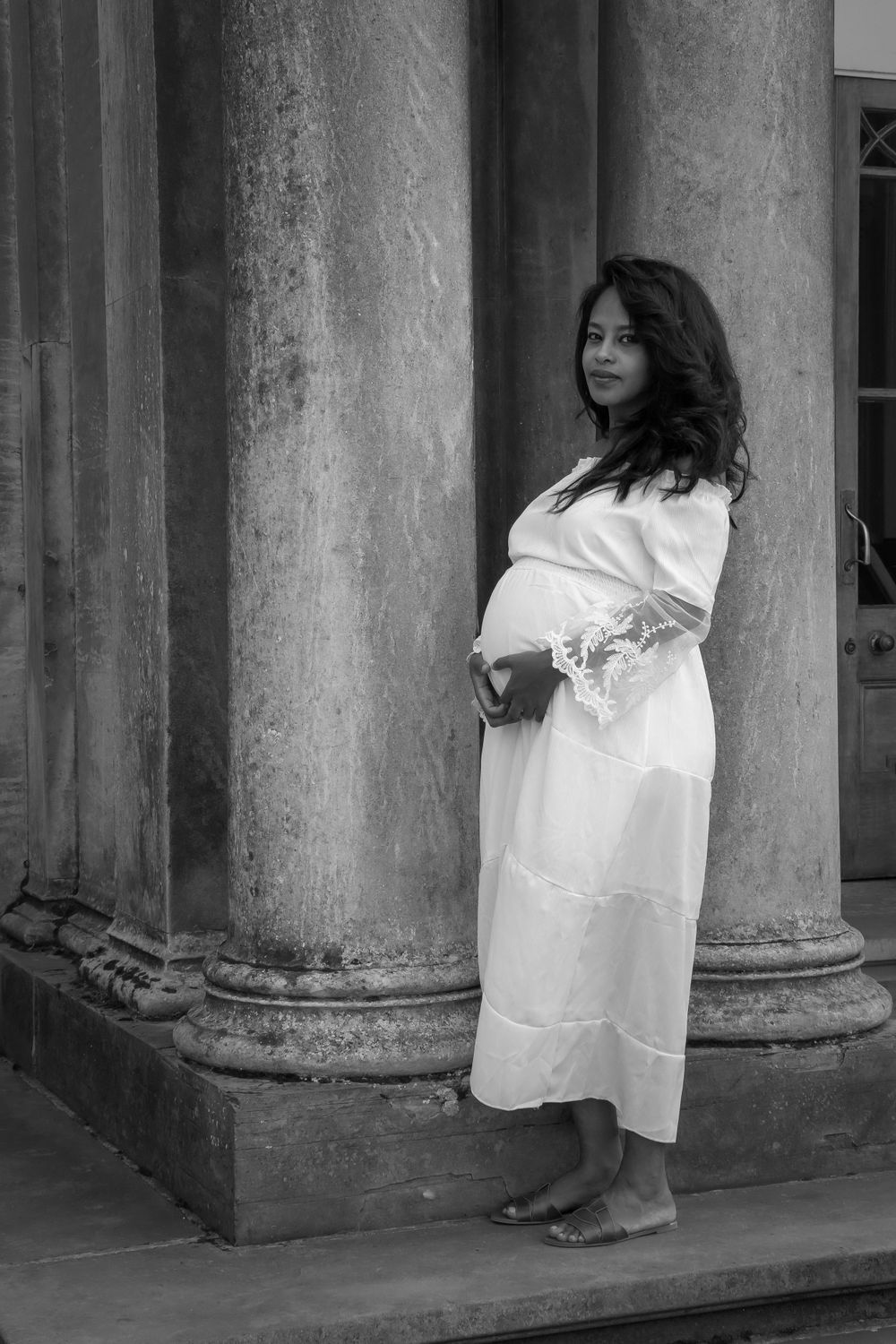 Black and white maternity photo of a pregnant woman in a white dress standing beside classical stone columns.