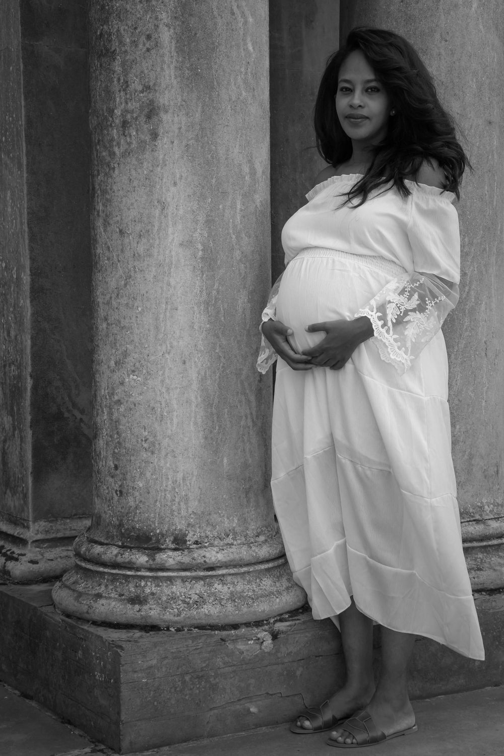 A pregnant woman in a flowing white dress poses against stone pillars in black and white photography.