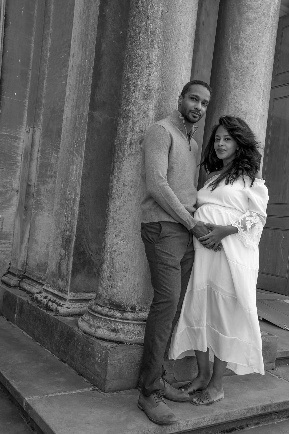 Couple embracing by stone columns in black and white, with the woman wearing a white maternity dress.