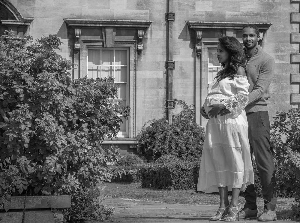 Tender moment between expectant parents outside a historic building with flowering bushes.