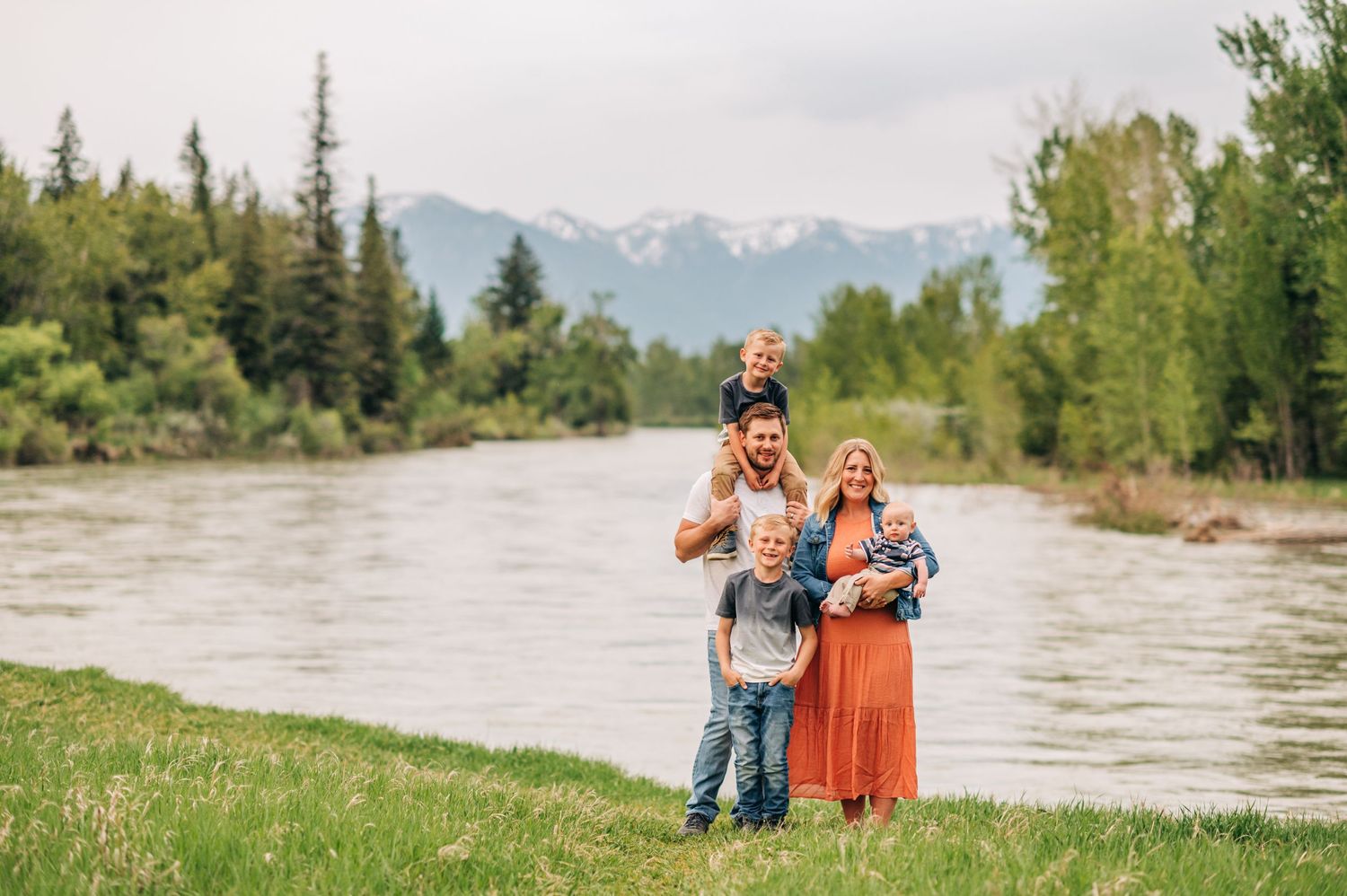 Family standing together by a river with snow-capped mountains in the background.