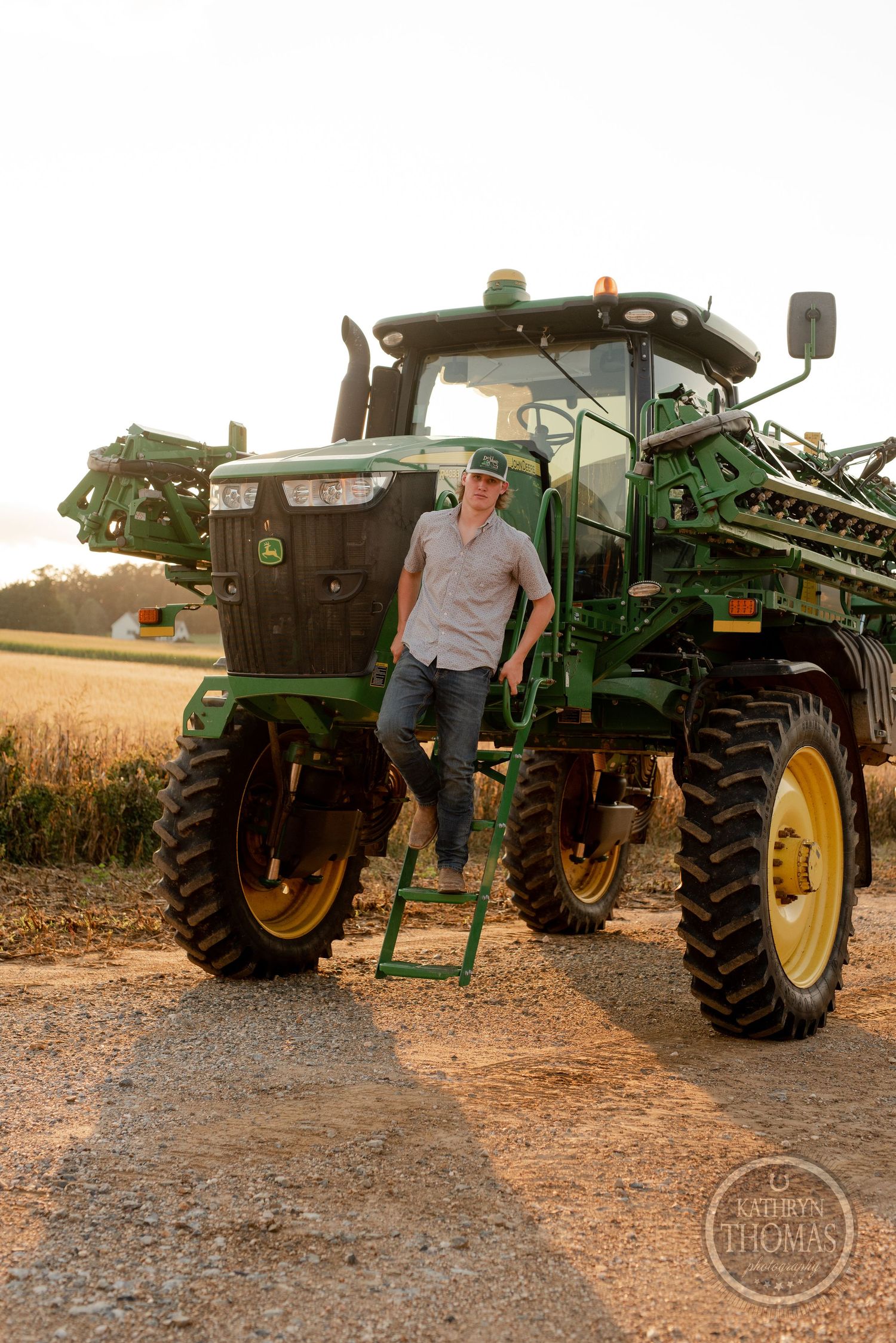 Farmer stands on the ladder of a large John Deere sprayer during sunset in a rural field.
