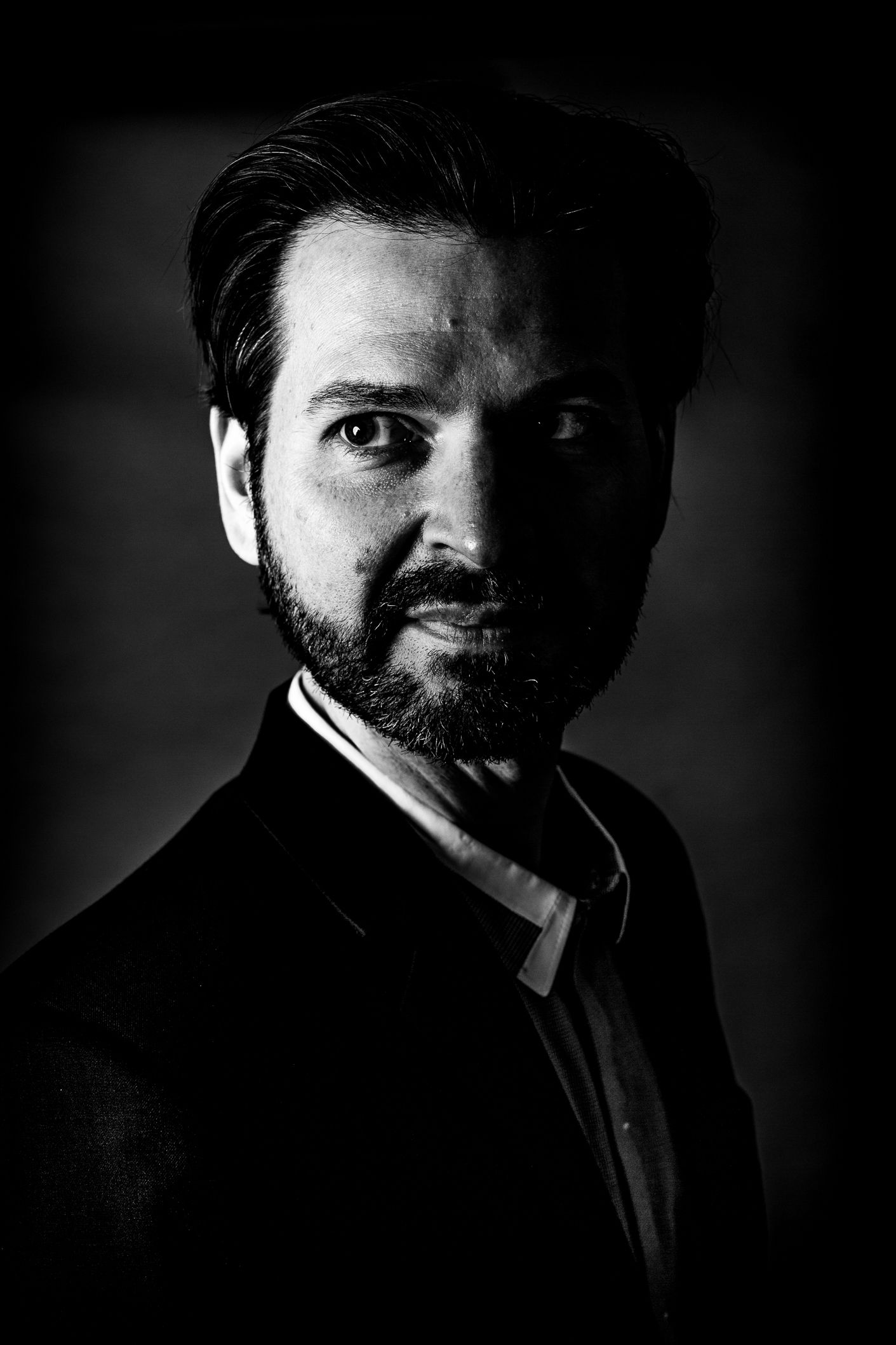 Dramatic black and white portrait of person in suit with beard against dark background.