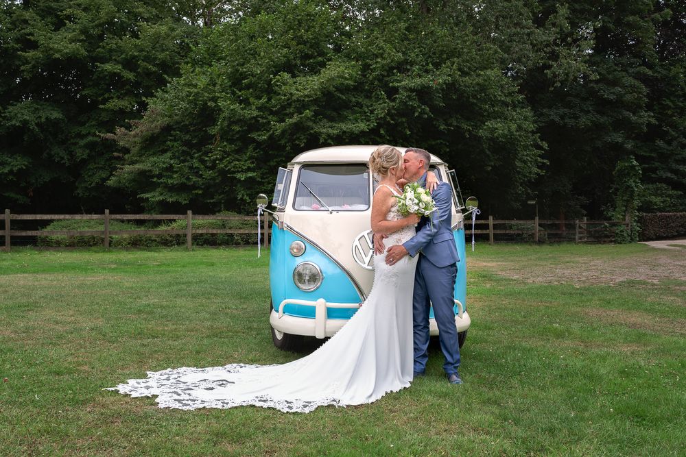 Classic blue and white VW camper van serves as backdrop for wedding photos on grassy field.