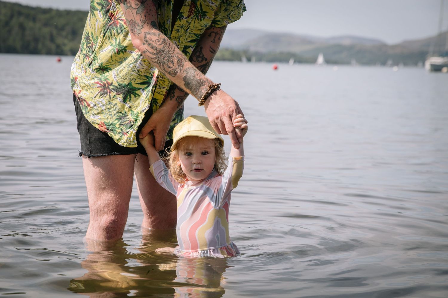 Adult holding childs hand while wading together in a lake with sailboats in the distance.
