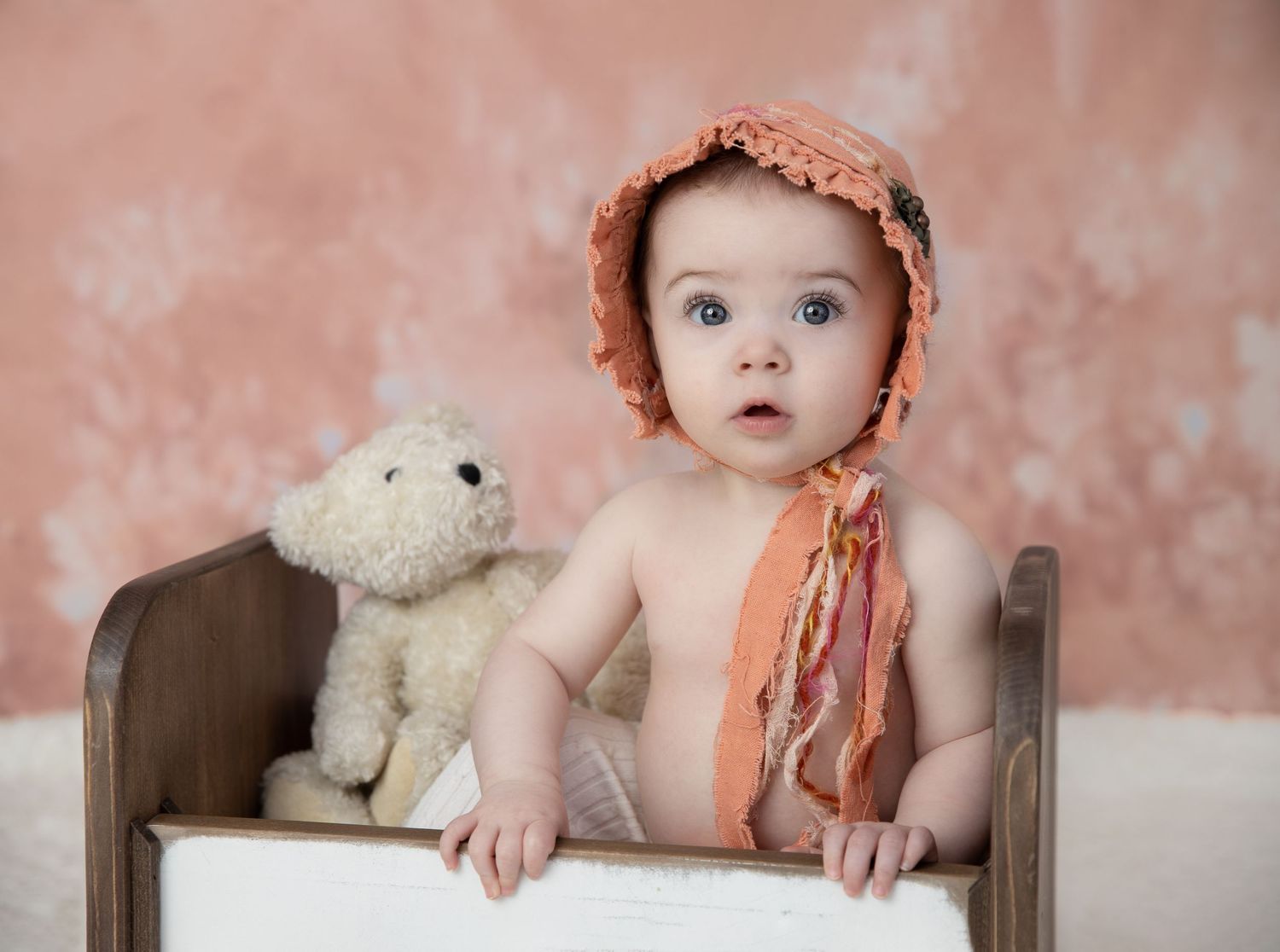 Baby wearing peach bonnet sitting with stuffed white teddy bear against pink backdrop.