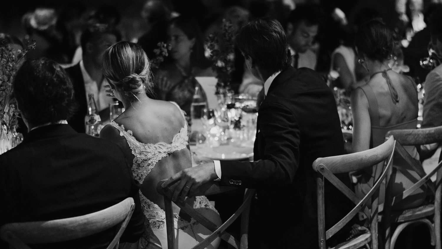Black and white photo captured at the wedding of Red & Camila. Couple are sat at the dinner table with guests.