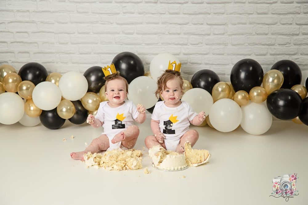 Twin babies sit on floor with smashed cakes and balloon garland behind them.