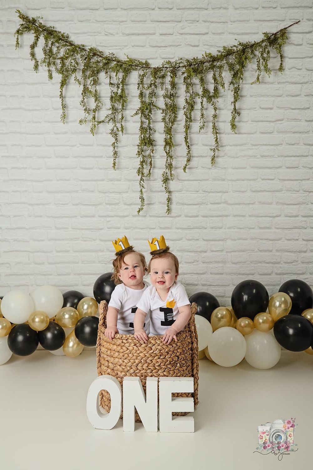 First birthday photo setup with greenery garland, balloon arch, and babies in a basket by letters spelling ONE.