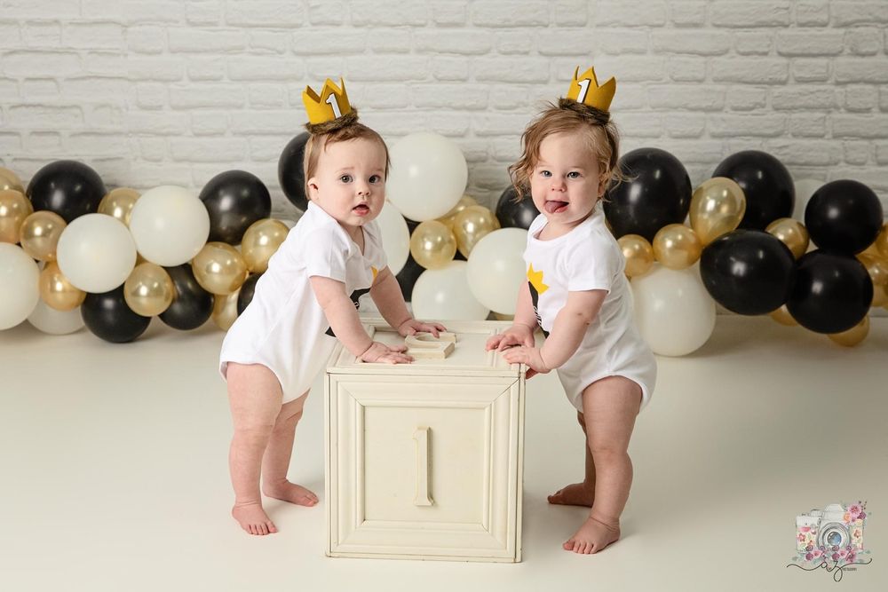 Babies in matching outfits stand beside decorative number one box with balloon backdrop.