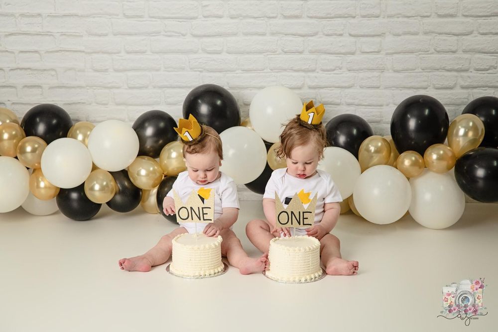 Twins in matching outfits sit with individual smash cakes surrounded by elegant balloon decor.