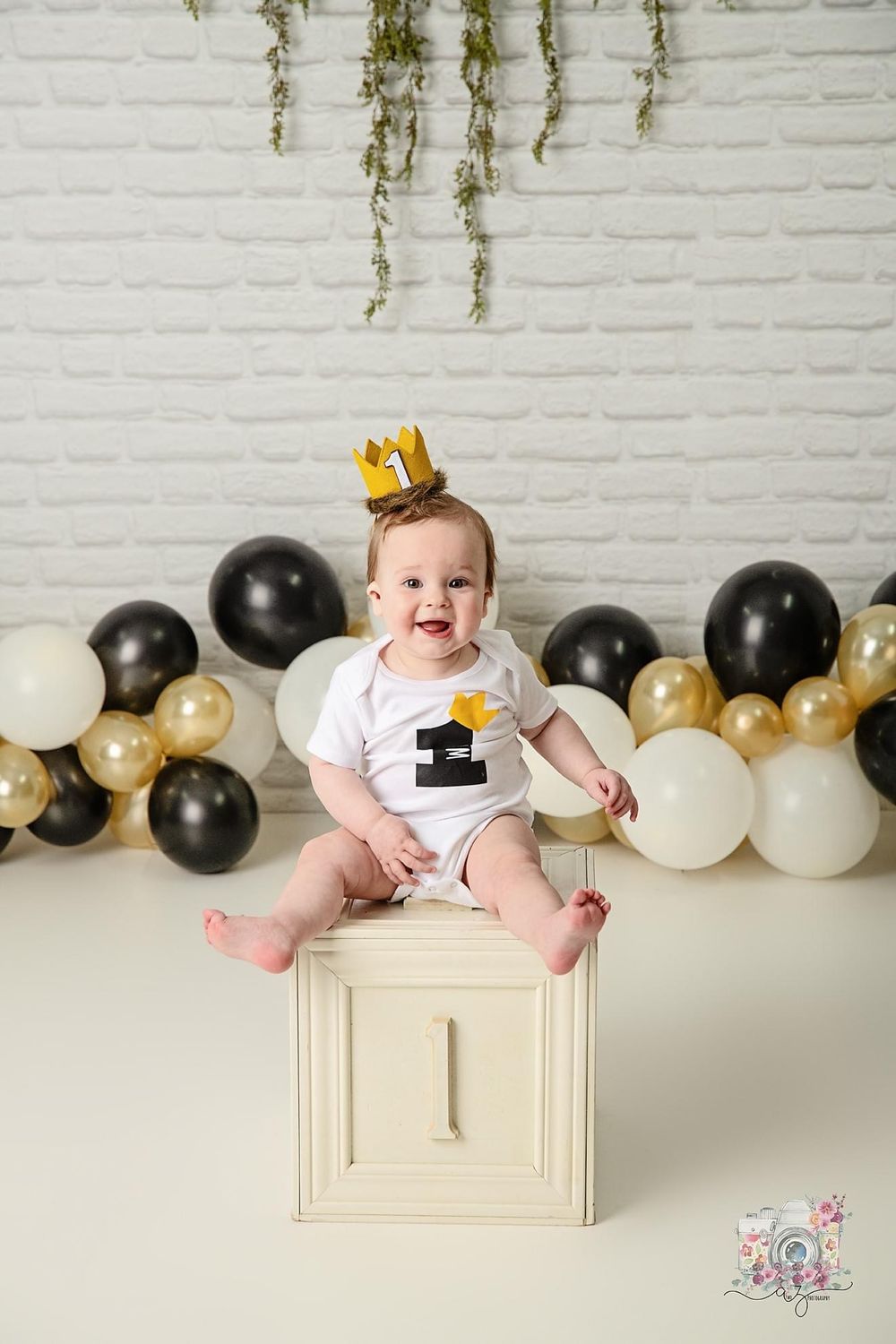 Baby in gold crown sits on decorative box with greenery and balloon backdrop.