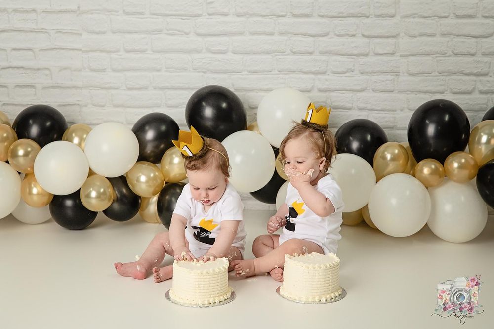 Two babies wearing gold crowns sit in front of black, white and gold balloon decorations with birthday cakes.