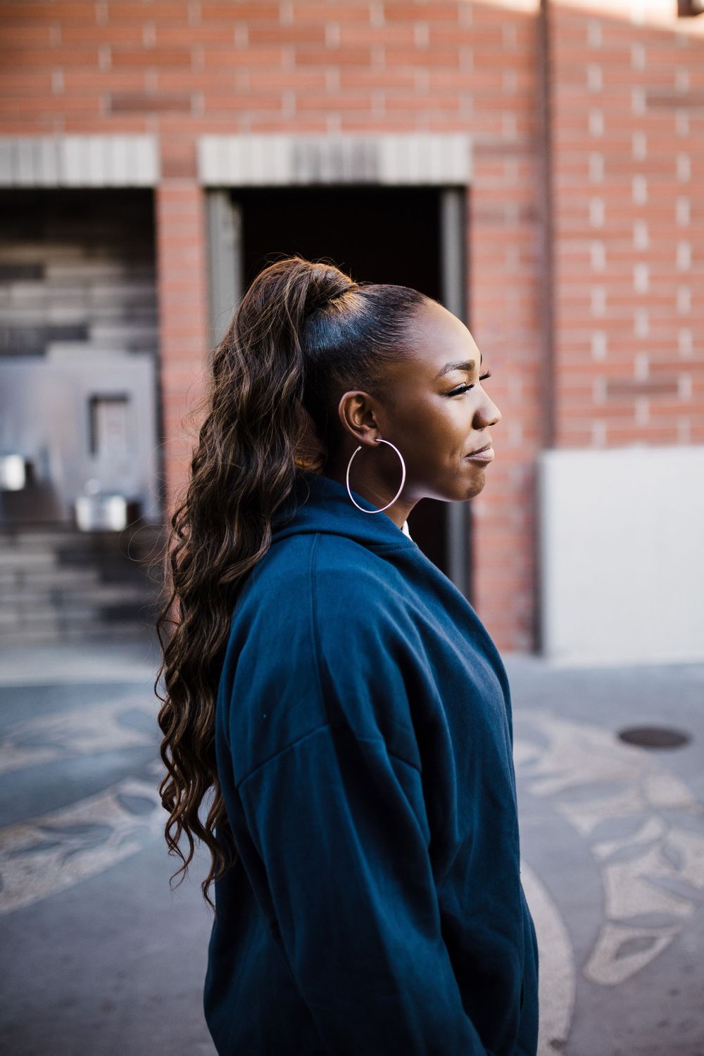 Young Black woman with long wavy ponytail and hoop earrings poses in profile outdoors near brick building.