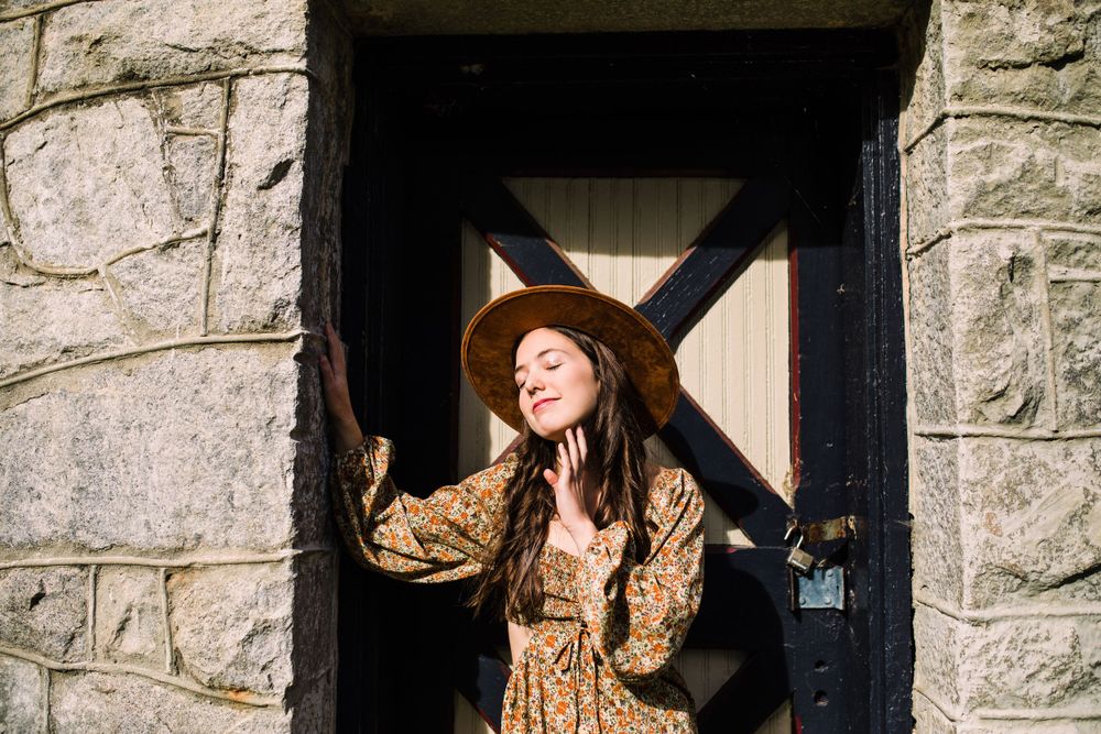 Young woman in floral dress and wide-brim hat standing in sunlit stone doorway with eyes closed.
