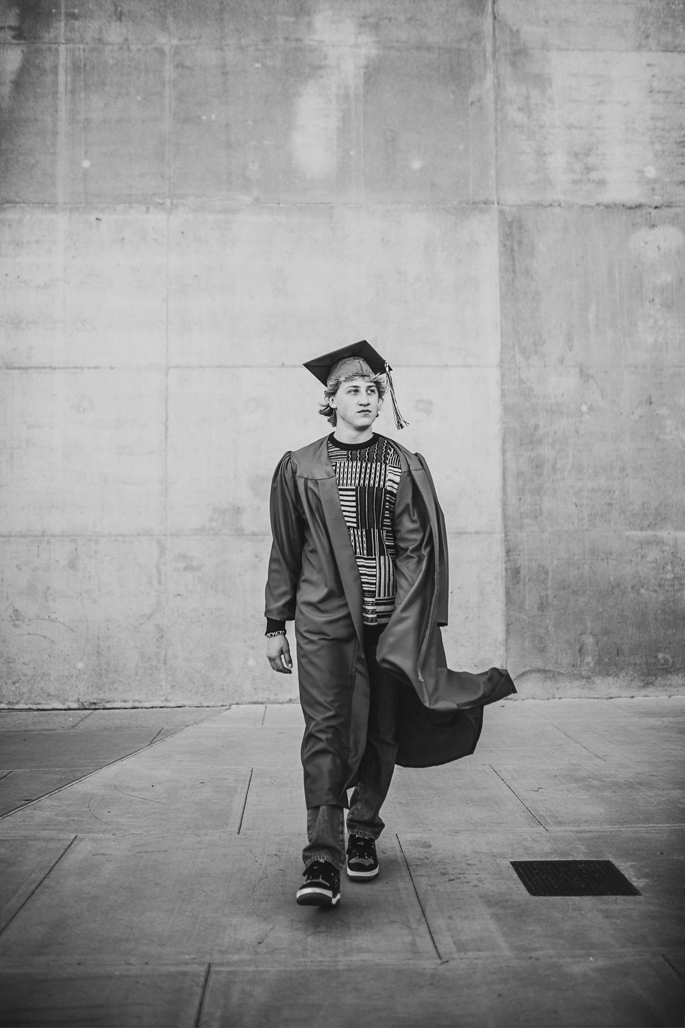 Black and white photo of young man walking in graduation cap and gown outdoors.