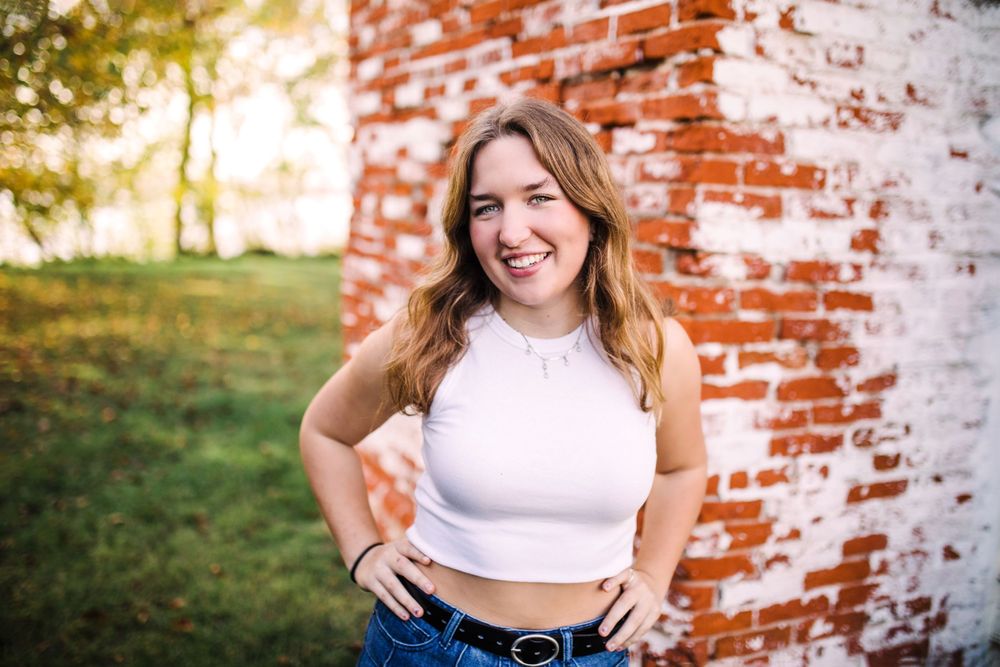 Smiling young woman in white crop top and jeans posing against a red brick wall outdoors.