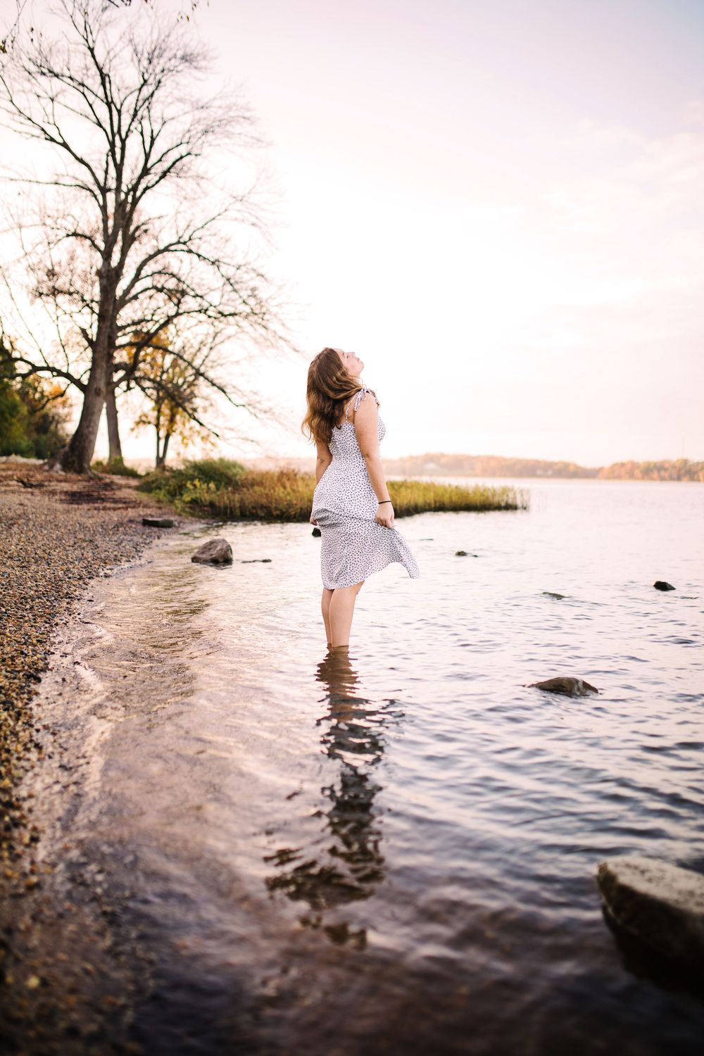 Young woman in white floral dress wading barefoot in calm lake water at golden sunset.