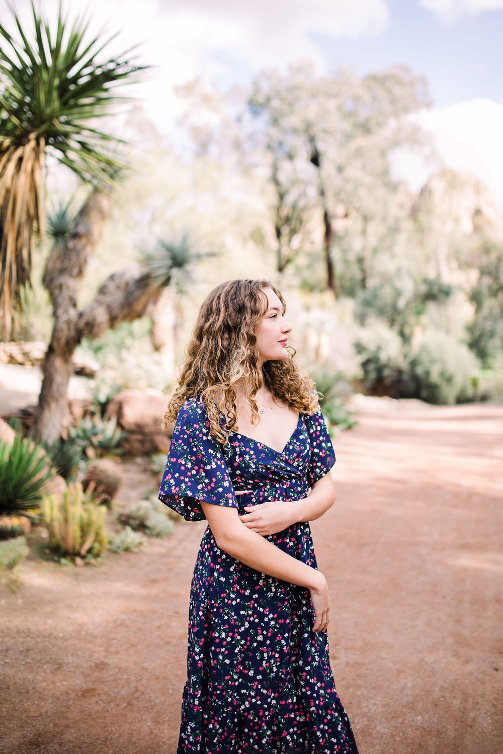 Young woman in navy floral dress standing in a desert garden with cacti and trees.