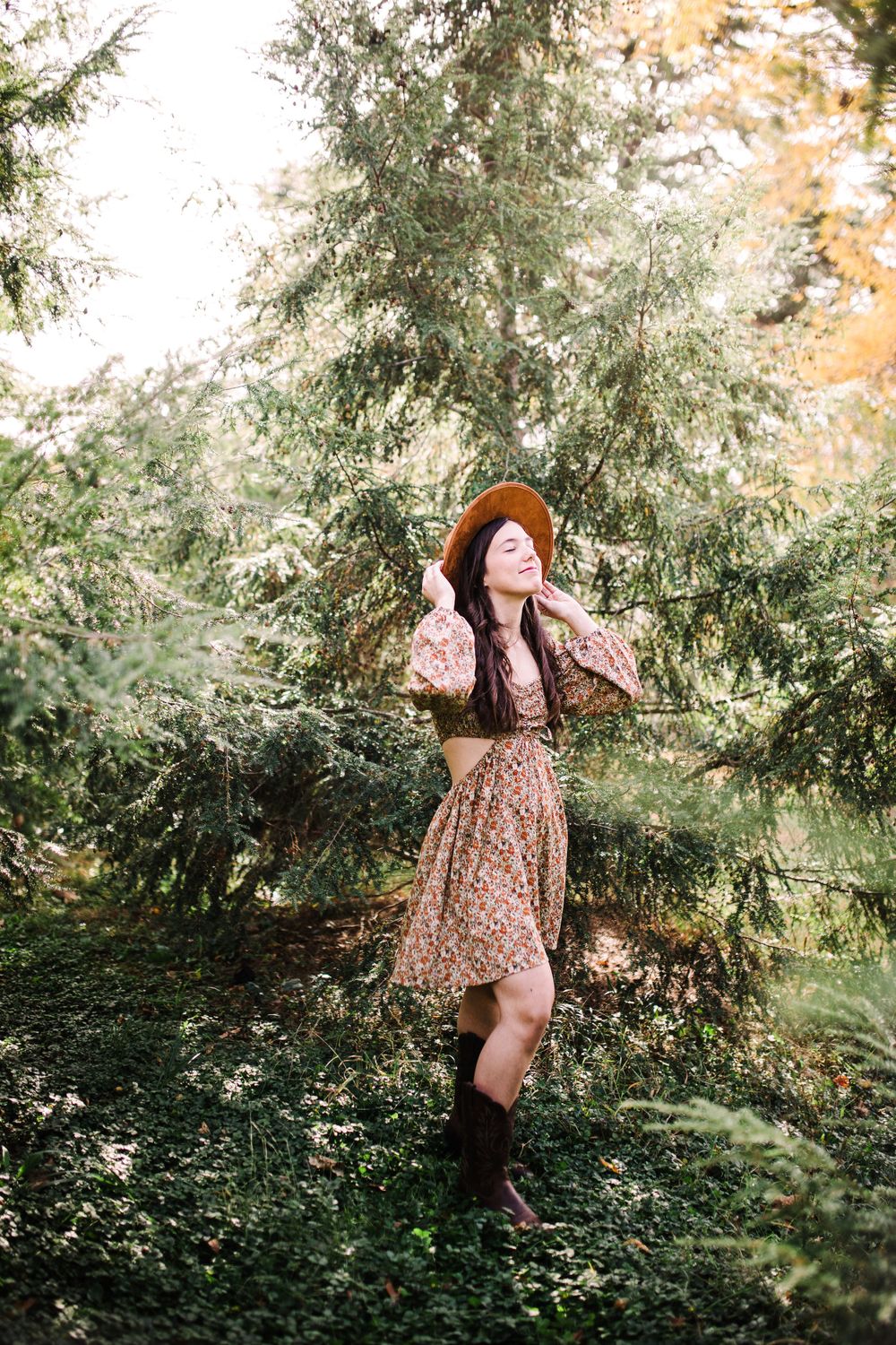 Woman in floral dress and brown wide-brim hat posing among evergreen trees in a sunlit forest.