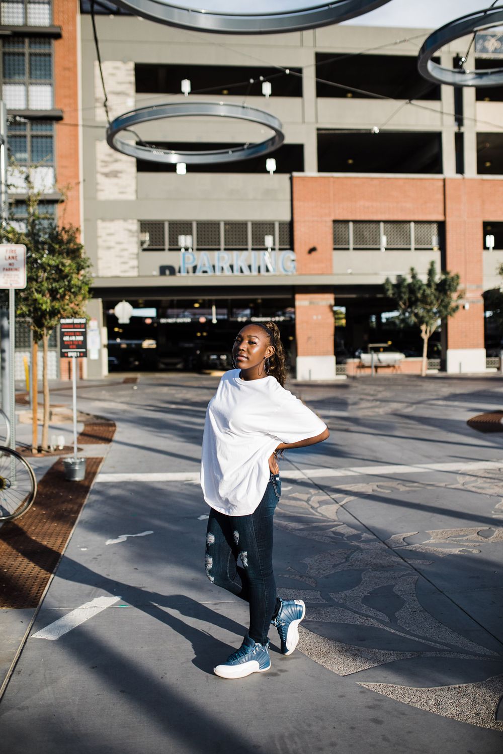 Young Black woman in white oversized tee and ripped jeans poses confidently in urban plaza setting.