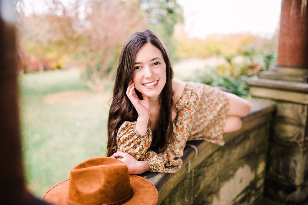 Smiling young woman with long dark hair leaning on stone wall holding brown hat in garden.