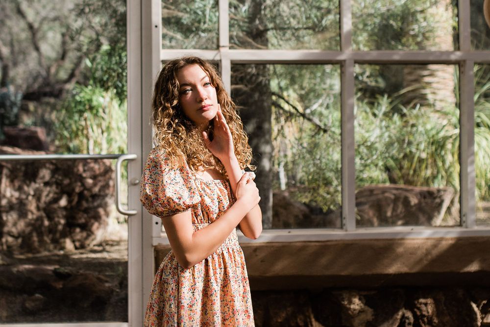 Young woman in floral dress posing by white window frame surrounded by lush greenery.