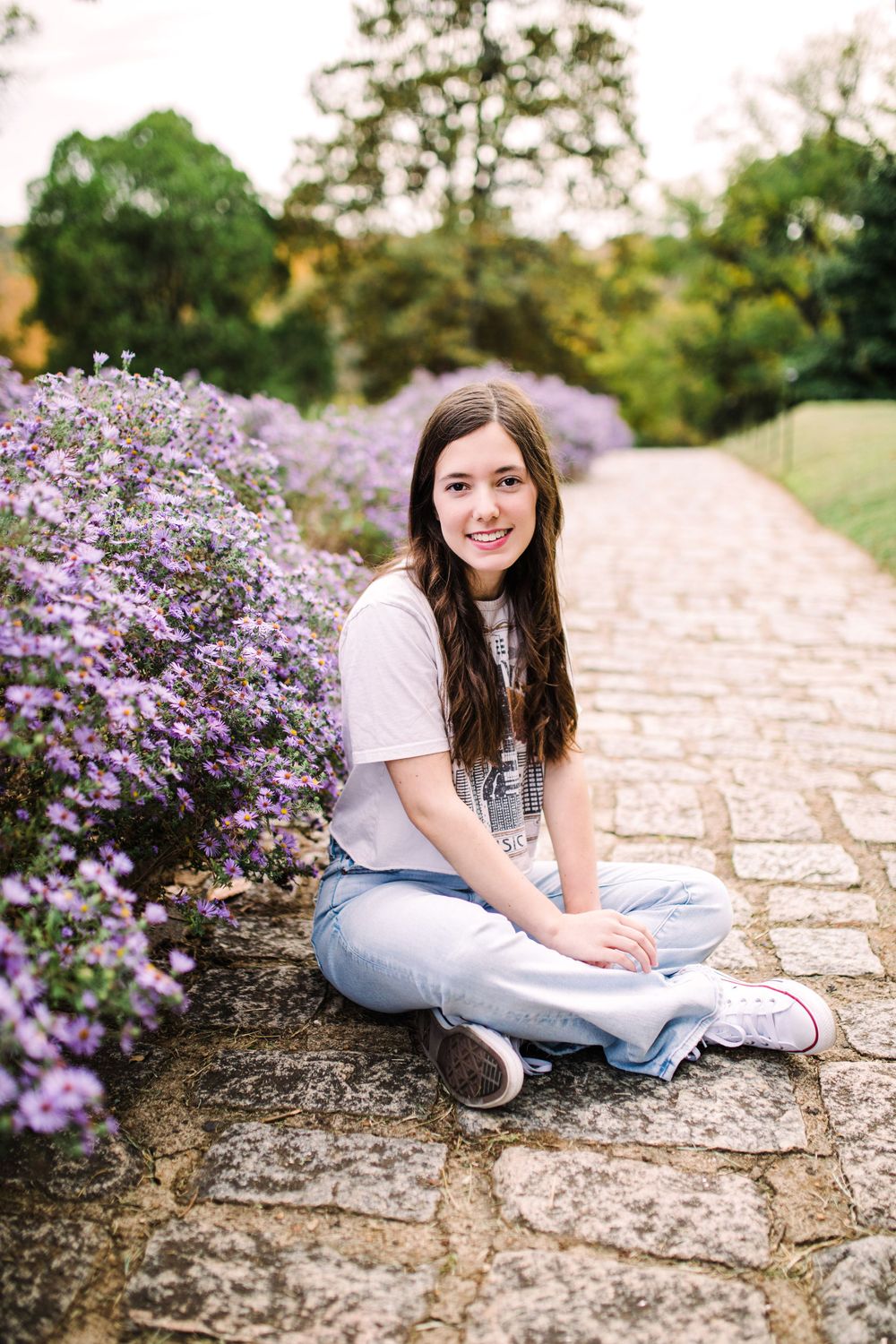 Teenage girl in white tee and jeans sitting cross-legged on cobblestone path beside purple flowers.