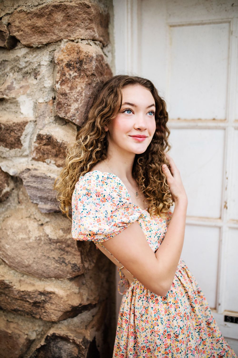 Young woman with curly hair in floral dress leaning against rustic stone wall outdoors.