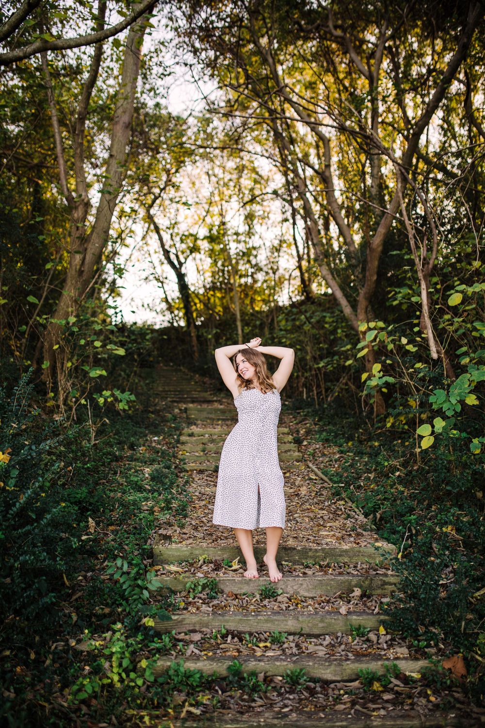 Young woman in white floral midi dress standing on moss-covered stone steps in a wooded forest path.