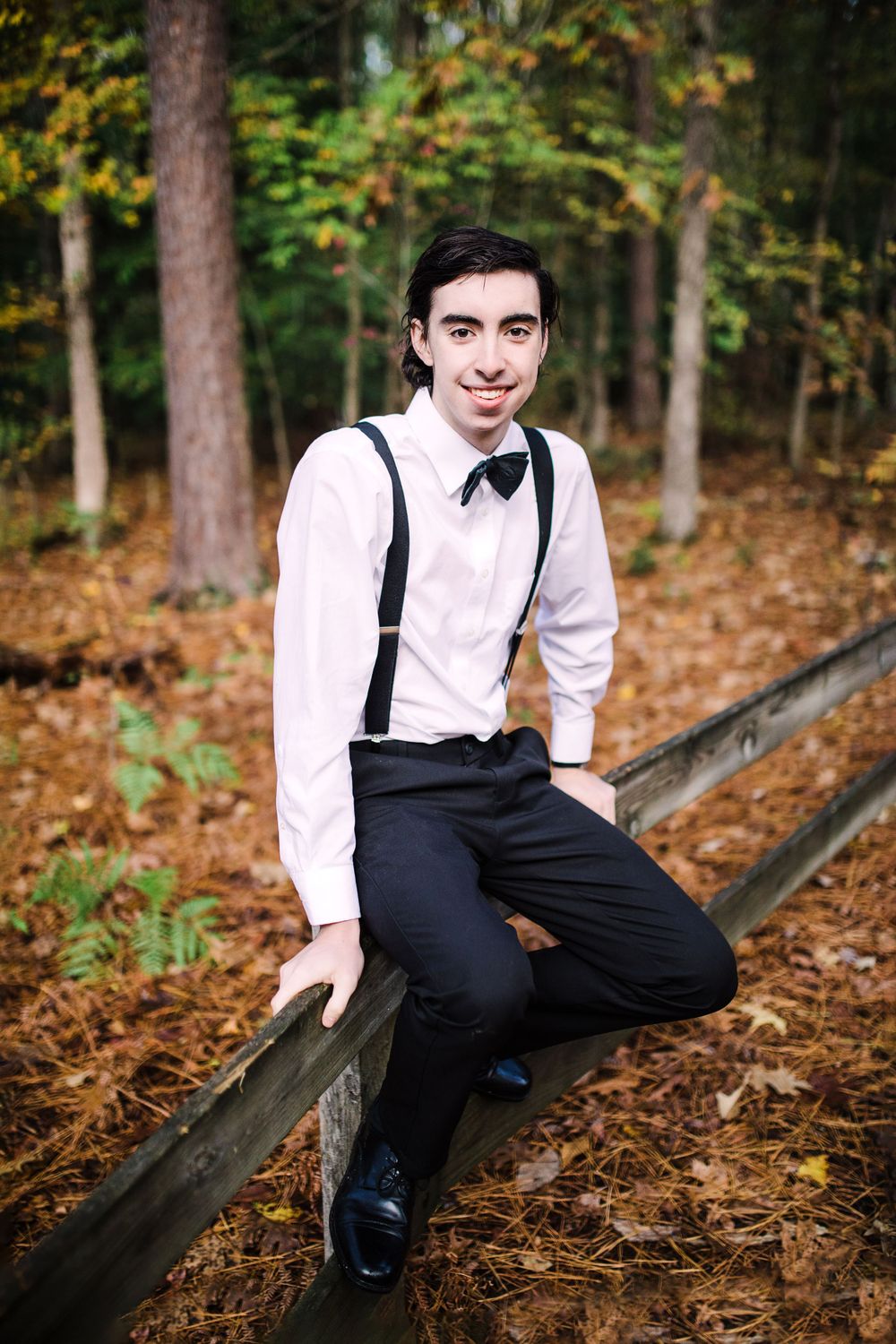 Young man in white shirt, suspenders, and bow tie sits on wooden fence in autumn forest setting.
