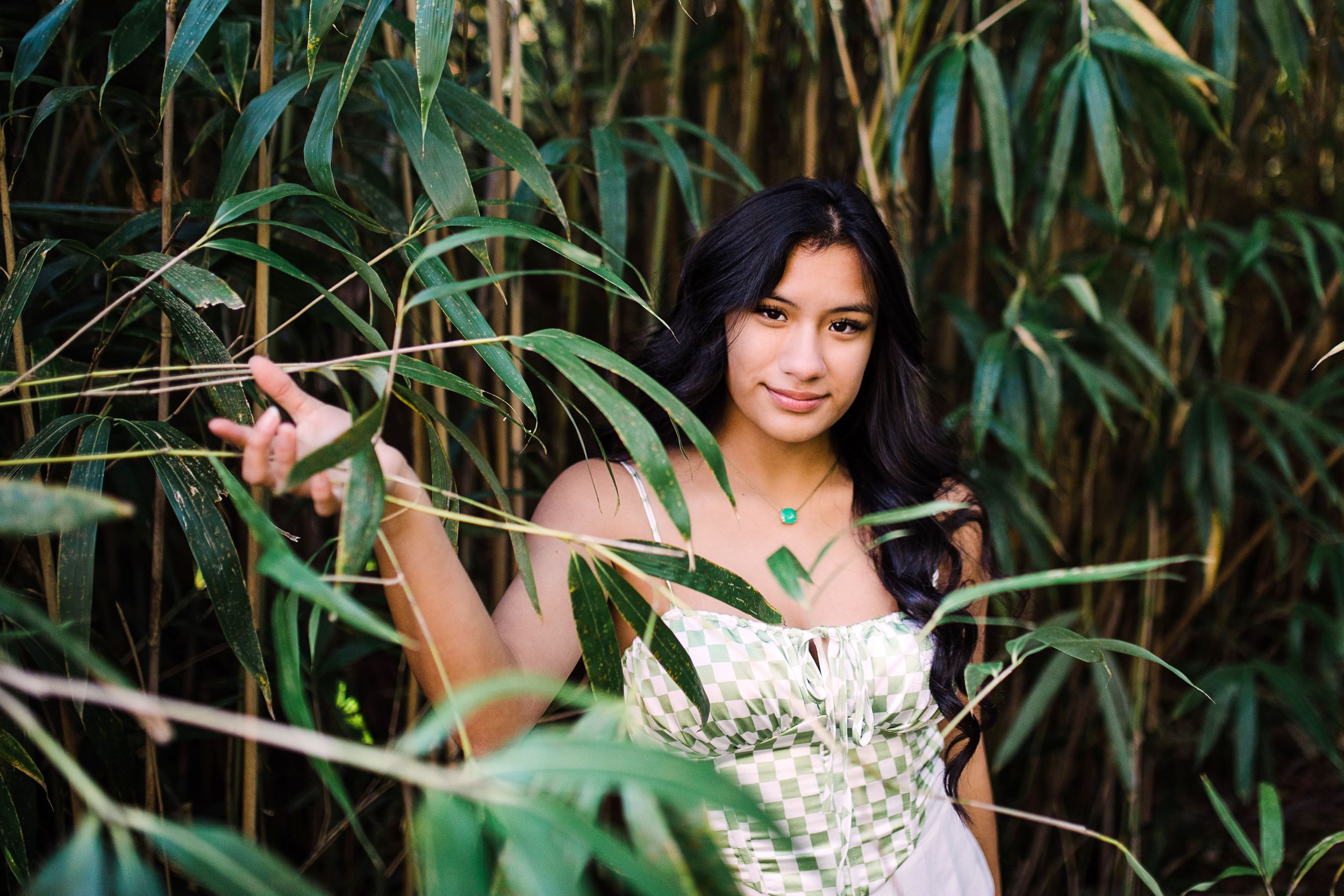 Young woman in green checkered top peers through bamboo leaves in a lush green garden.