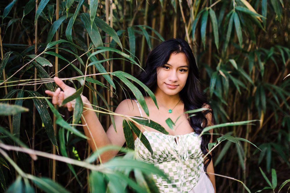 Young woman in green checkered top peers through bamboo leaves in a lush green garden.