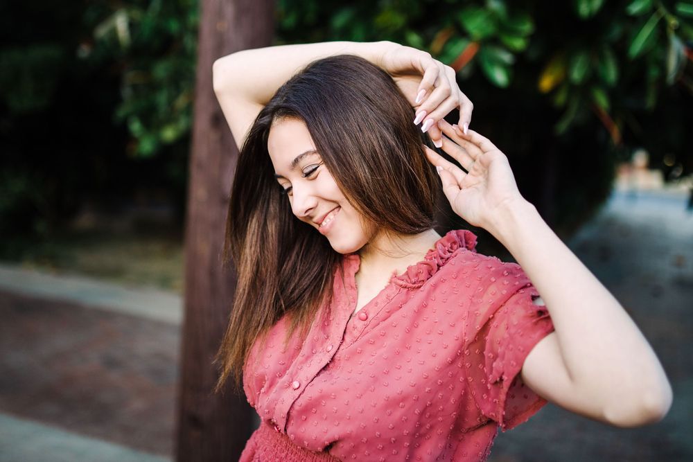 Smiling brunette woman in red top posing outdoors near green foliage in warm light.