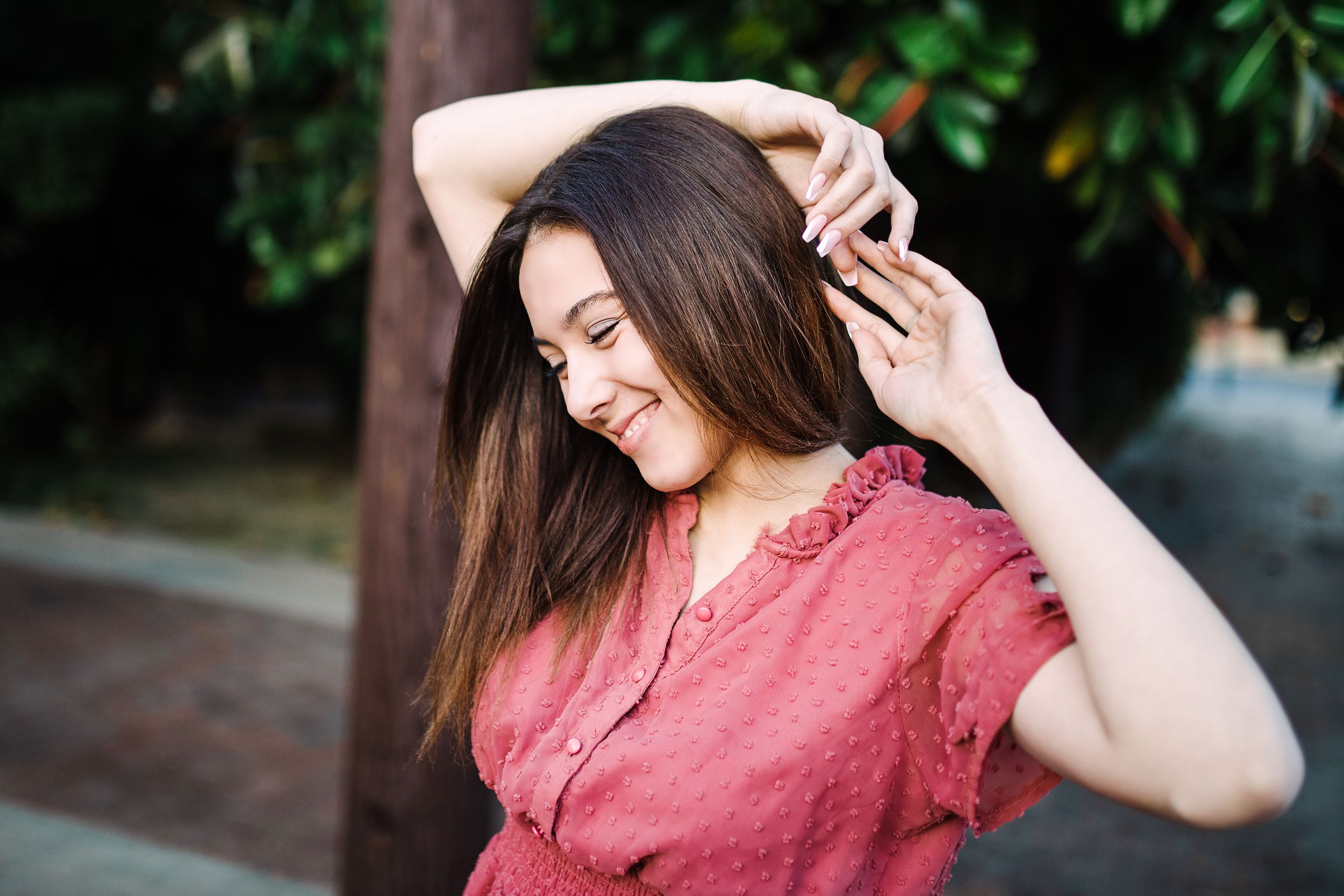 Smiling brunette woman in red top posing outdoors near green foliage in warm light.