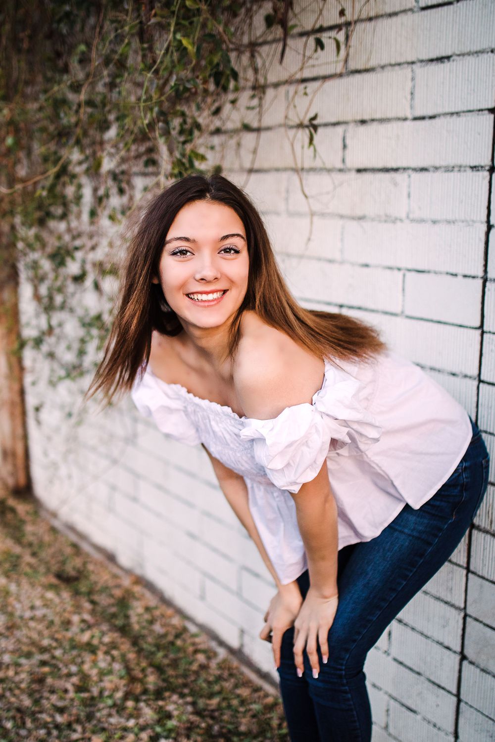 Smiling young woman in white off-shoulder top leaning against white brick wall outdoors.