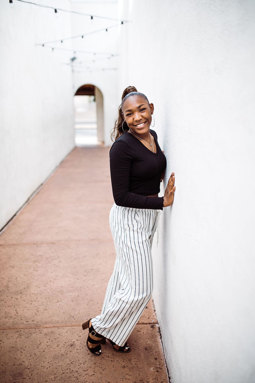 Smiling young Black woman in black top and striped wide-leg pants leans against white wall in corridor.