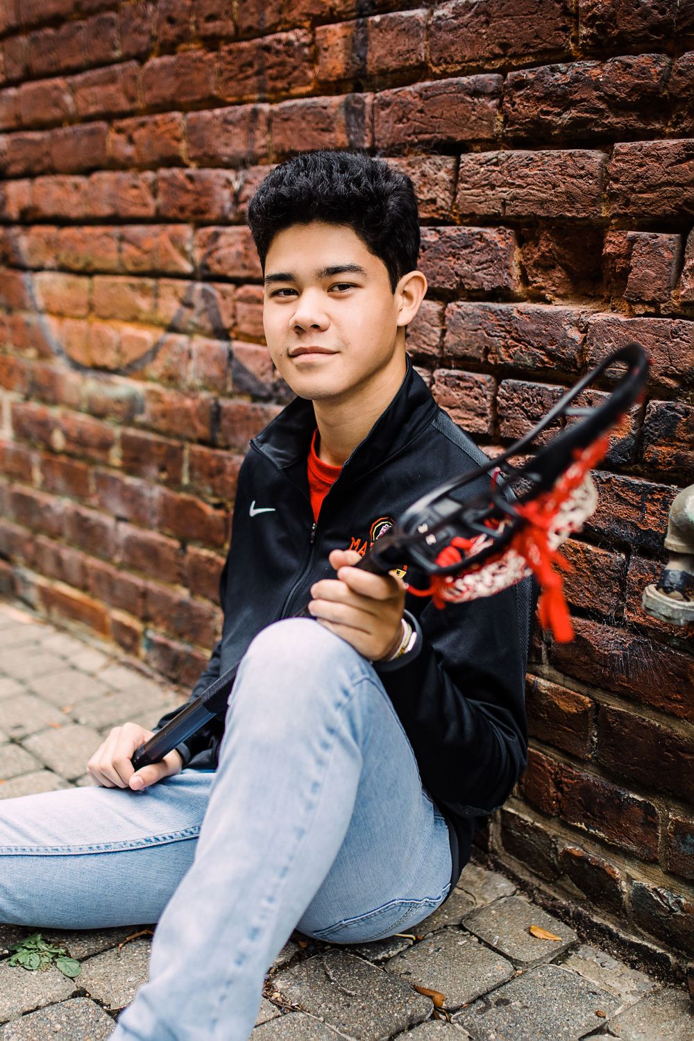 Young man sitting against brick wall holding lacrosse stick wearing Nike jacket and jeans.