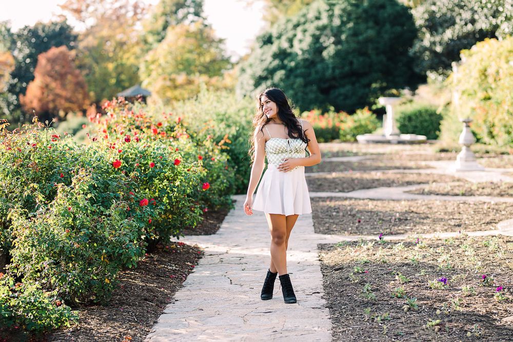 Young woman in white skirt and green top walks along a rose garden pathway in warm autumn sunlight.