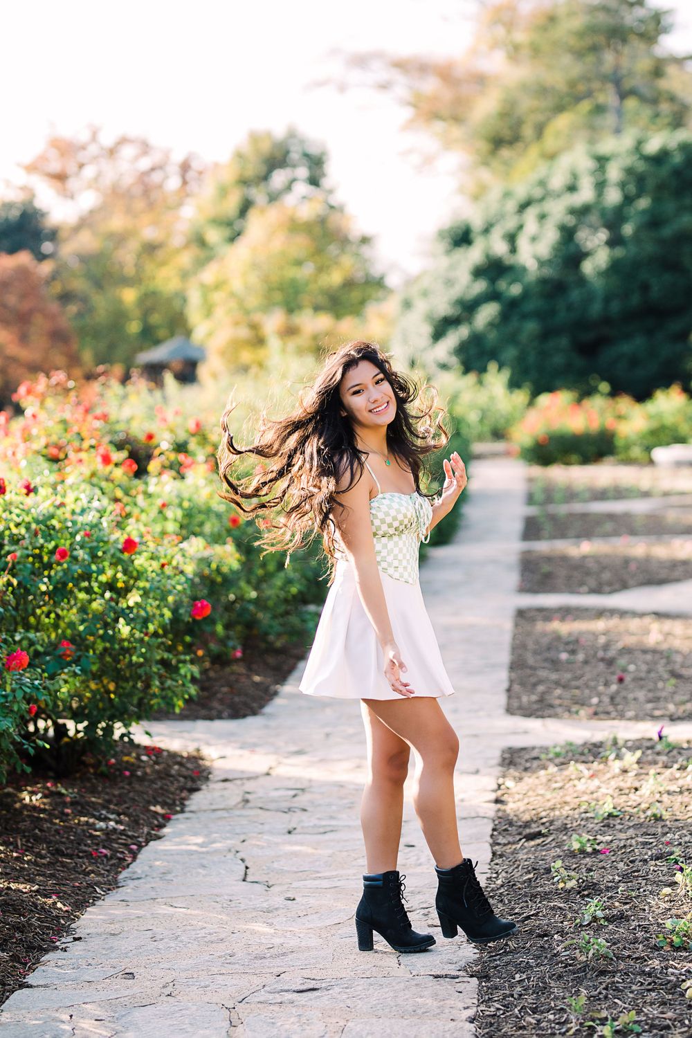 Joyful young woman in white dress and black boots twirls on a garden path lined with colorful roses.