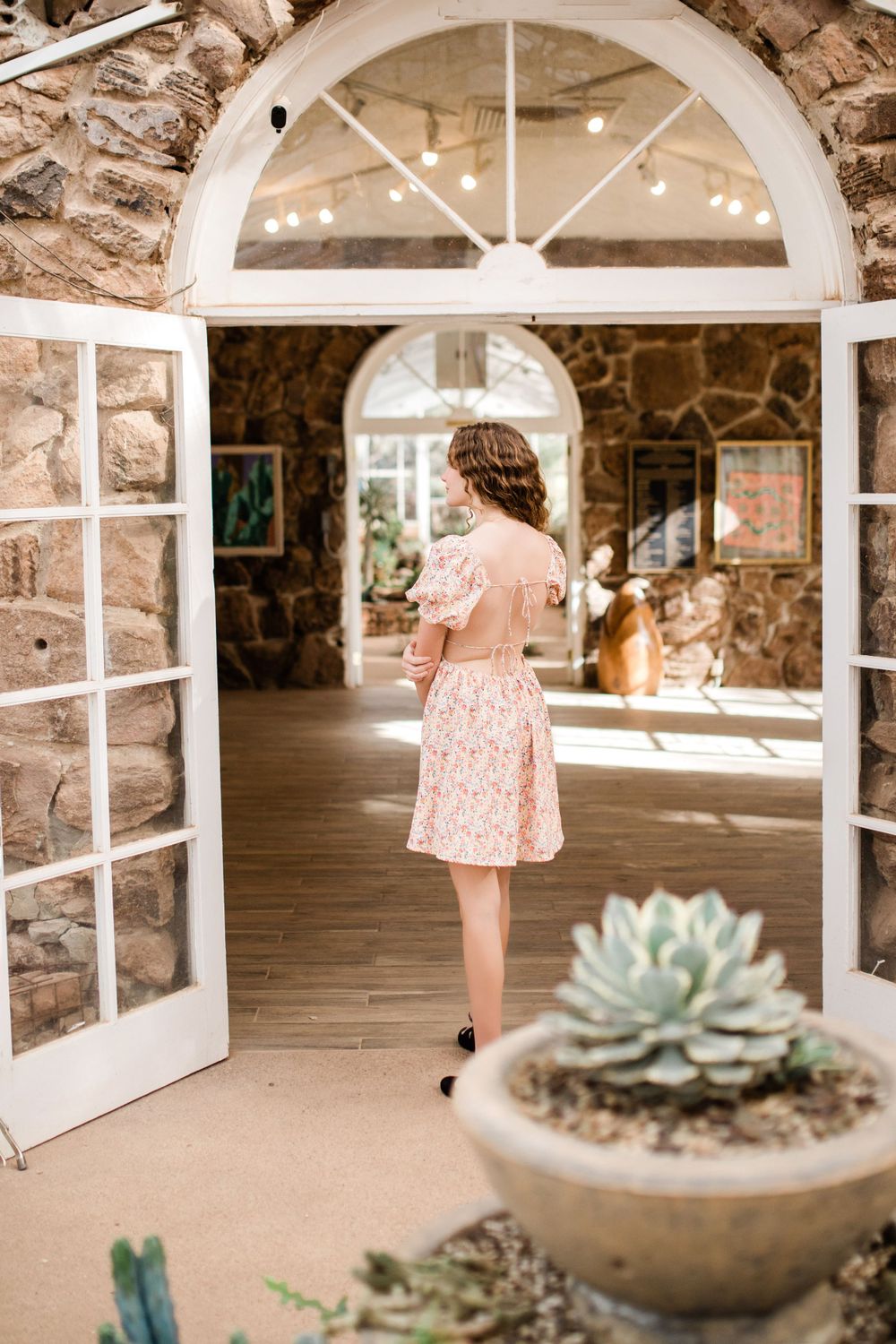 Young woman in floral dress walking through arched white doors of a stone building.